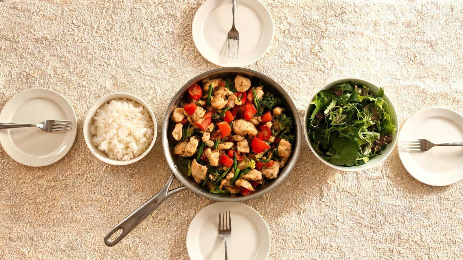 Overhead view of a skillet of chicken, broccoli, red pepper and asparagus beside a bowl of white rice, a bowl of green salad, and four empty white plates with forks on a beige table. Illustrative image.